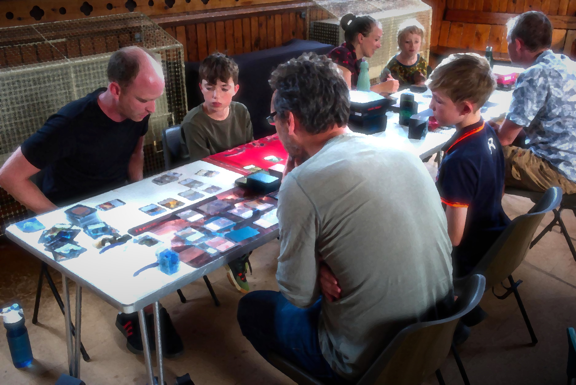 a group of adults and children sat at tables playing Magic the Gathering